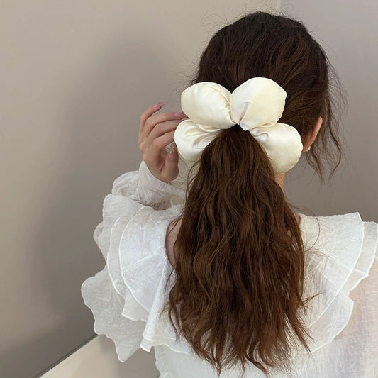 Woman with long brown hair tied in a ponytail with a large cream-colored scrunchie against a neutral background.